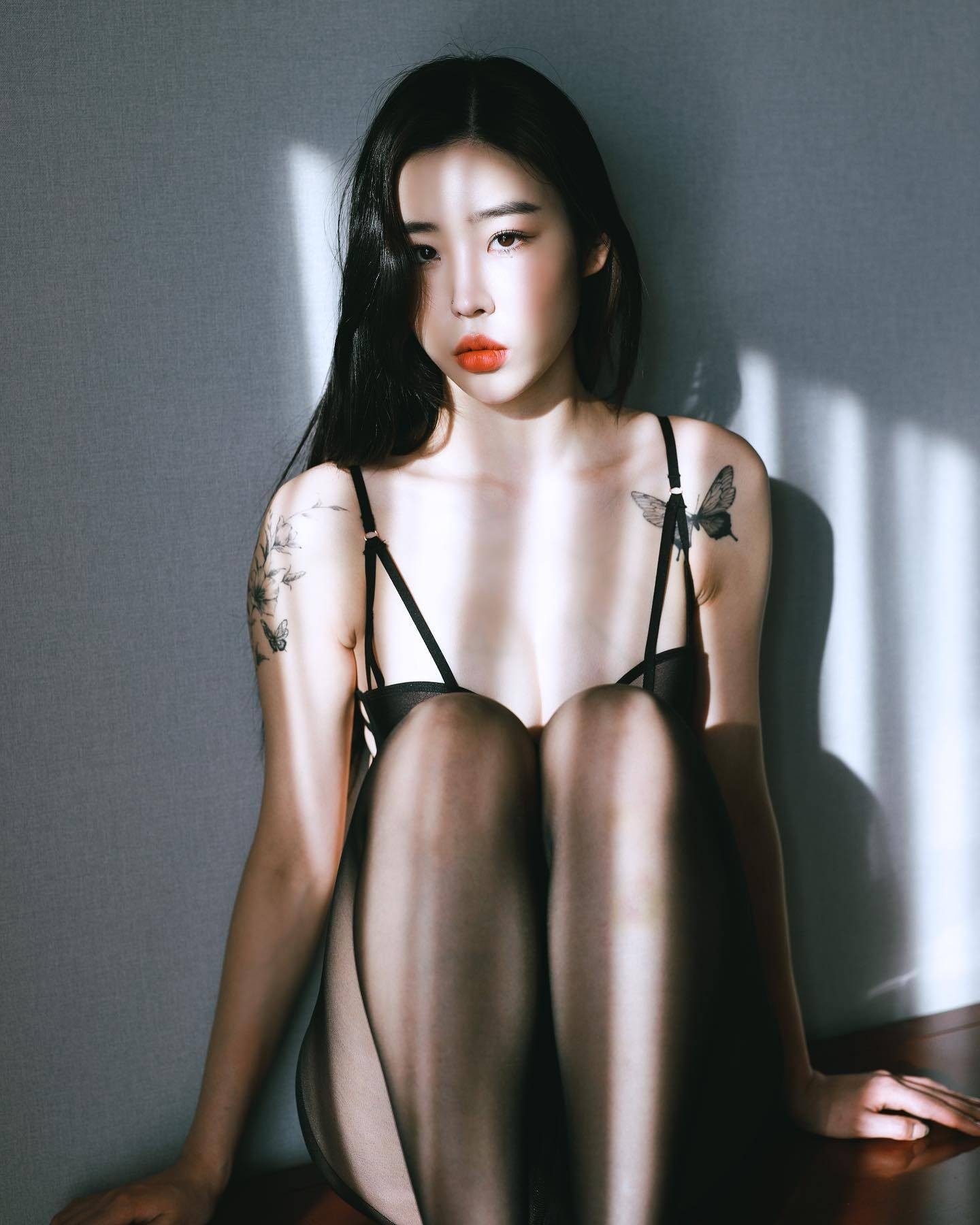 整体造型完整展示，模特倚靠墙壁，黑色内衣、丝袜与红唇构成经典对比，纹身成为最独特的配饰
