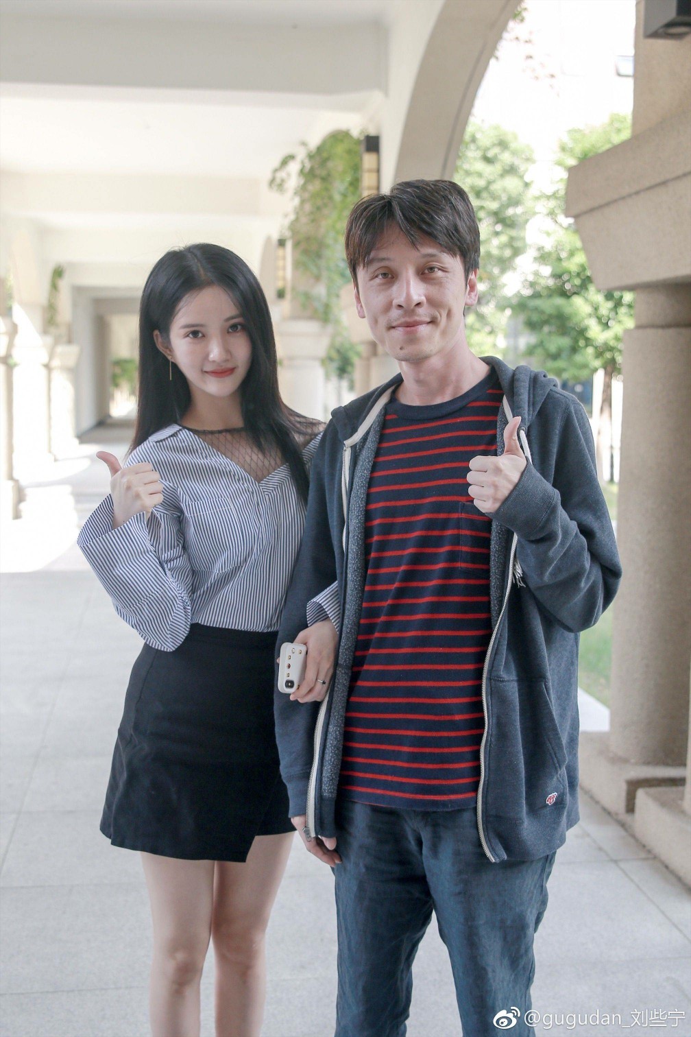 刘些宁身着黑色短裙与长靴侧身倚靠灰墙，都市光影勾勒出其优雅的身形轮廓